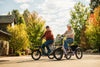 Two helmeted riders on electric tricycles along a street with autumn trees
